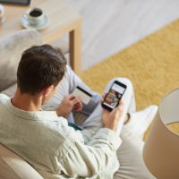 Rear view of young man sitting on sofa with mobile phone and shopping online using his credit card